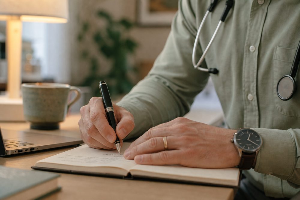 Clinician writing careful patient notes beside a laptop